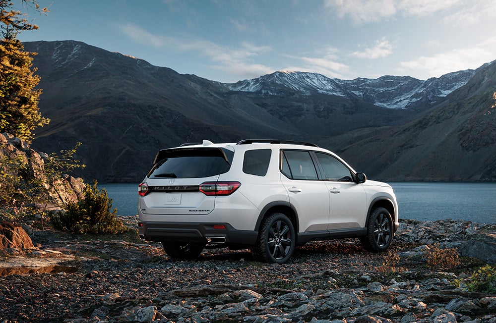 White Honda SUV parked on a rocky mountain landscape at sunset, with snow-capped peaks and evergreen trees in the background.