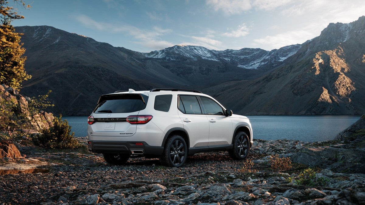 Rear three-quarter view of a white 2025 Honda Pilot Black Edition SUV parked on a rocky shore overlooking a lake and snow-capped mountains.
