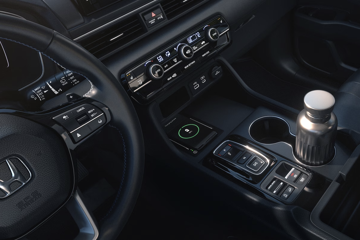 Close-up of the center console area of a Honda vehicle, showing the gear selector buttons, electronic parking brake, automatic brake hold, window controls, wireless charging pad with a phone charging, cup holders with a stainless steel water bottle, and climate control buttons.