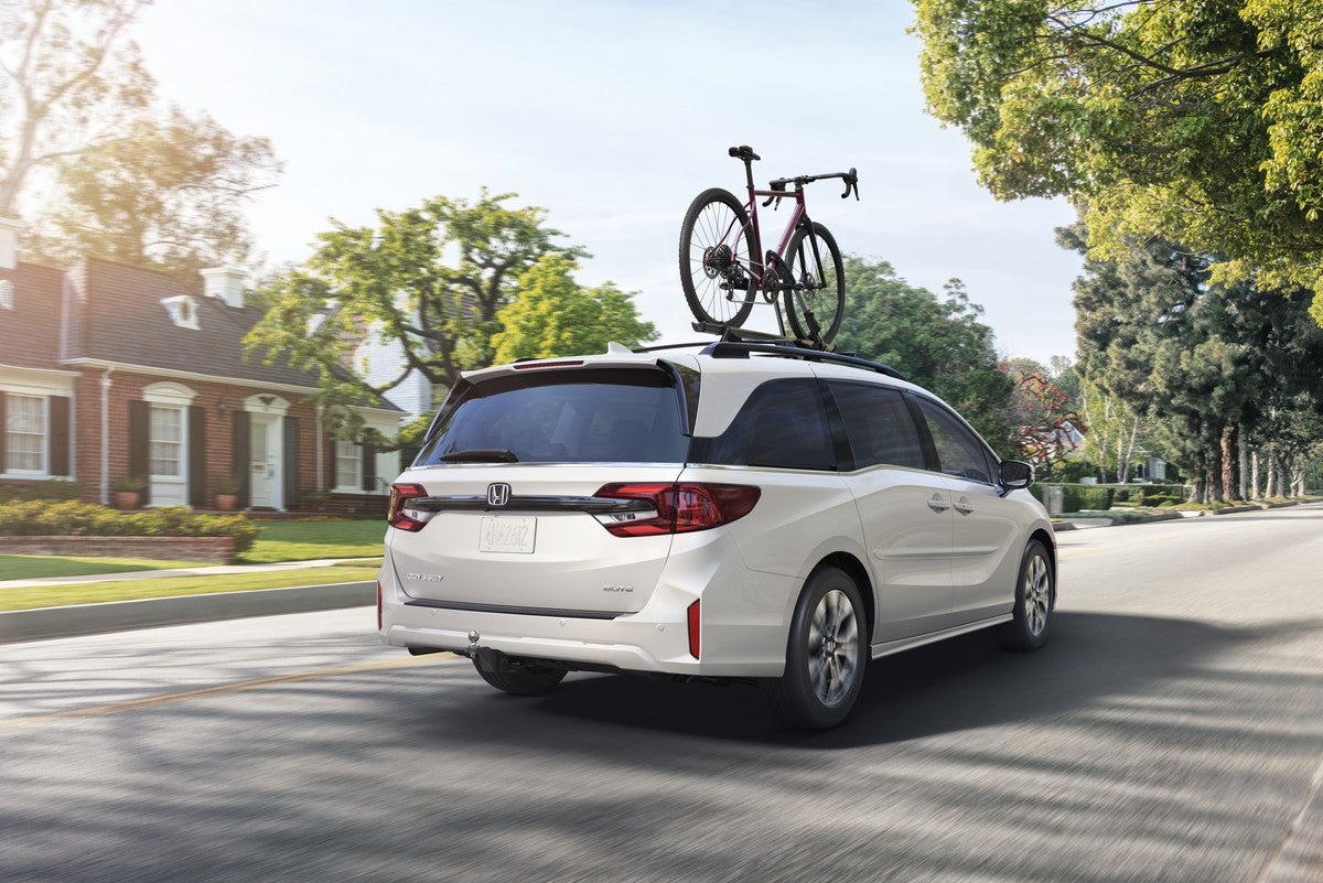 A white new Odyssey in Phoenix with a bicycle attached to the roof drives away from the viewer to the right on a residential suburban street.