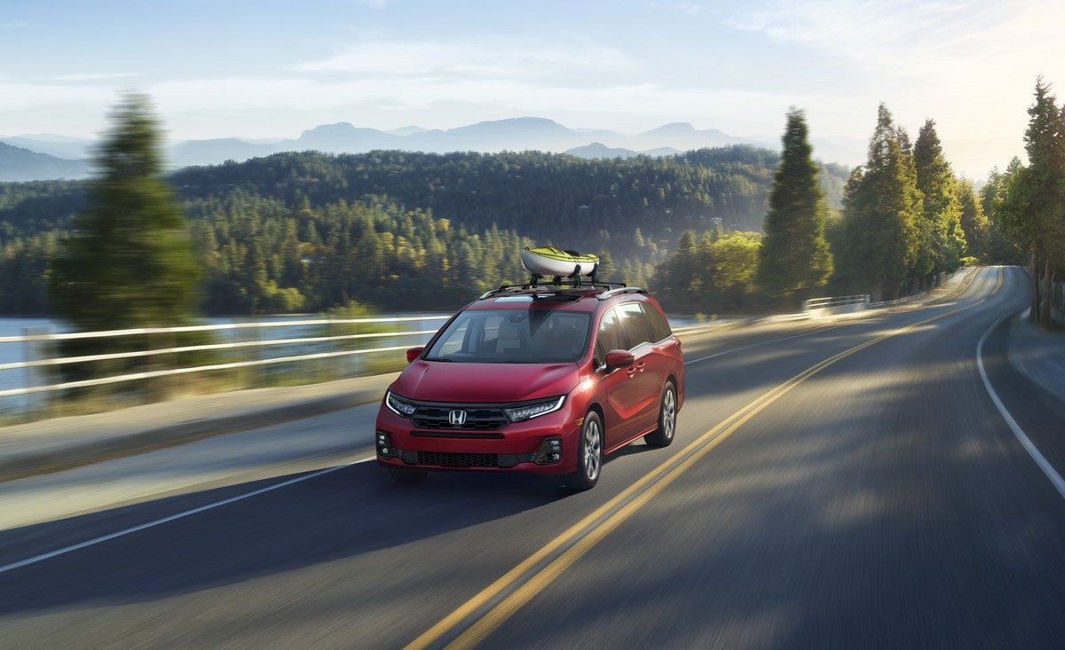 A red 2026 Odyssey in Phoenix with a green and white canoe attached to its roof drives towards the viewer on a long rural two-lane road through forested hills with a small lake to the left.