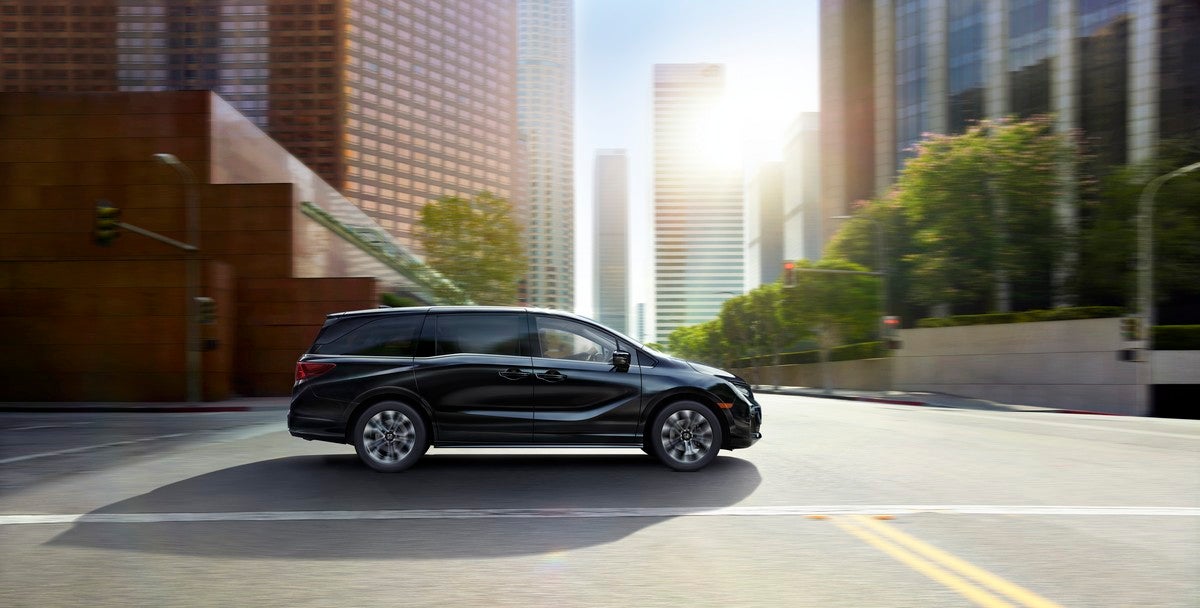 A black 2026 Honda Odyssey in Avondale drives to the right on a city street through an intersection with light shining from behind the vehicle.