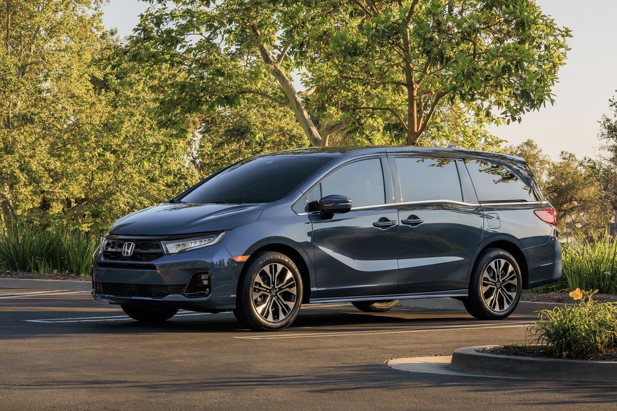 A dark blue 2026 Honda Odyssey in Phoenix is parked facing the viewer to the left in a small parking lot with grass and small trees in the background.