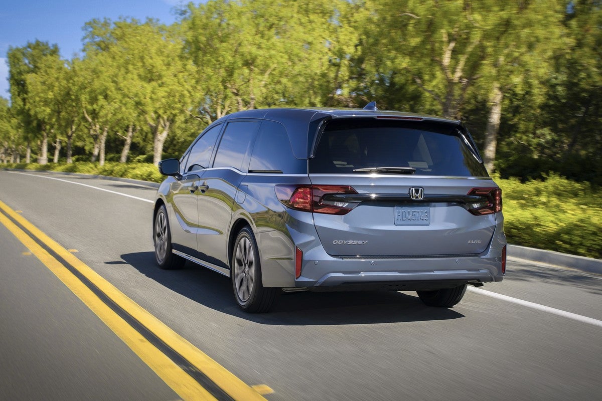 A light blue new Honda Odyssey in Avondale drives away from the viewer to the left on a rural two-lane road with a forest in the background.