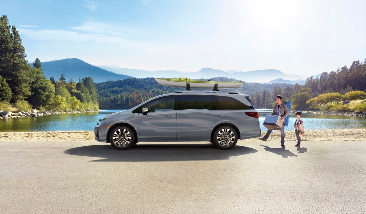 A silver new 2026 Honda Odyssey in Phoenix is parked facing left in front of a lake and a distant forest and mountains. A father and son load camping gear into the vehicle, which has a kayak attached to the roof.