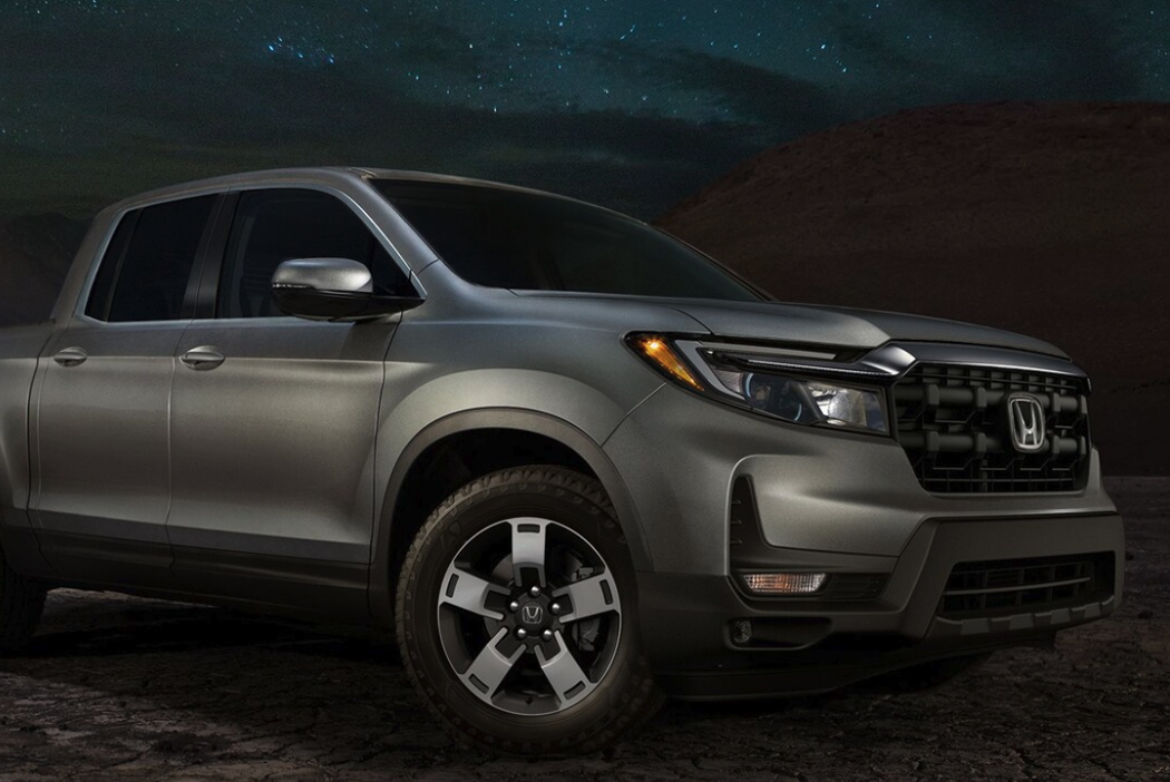 A gray Honda Ridgeline pickup truck is shown in a low-light, close-up shot of its front corner, highlighting the grille, headlamp, and wheel. The background is a dark night sky with stars over a rugged, dark terrain.
