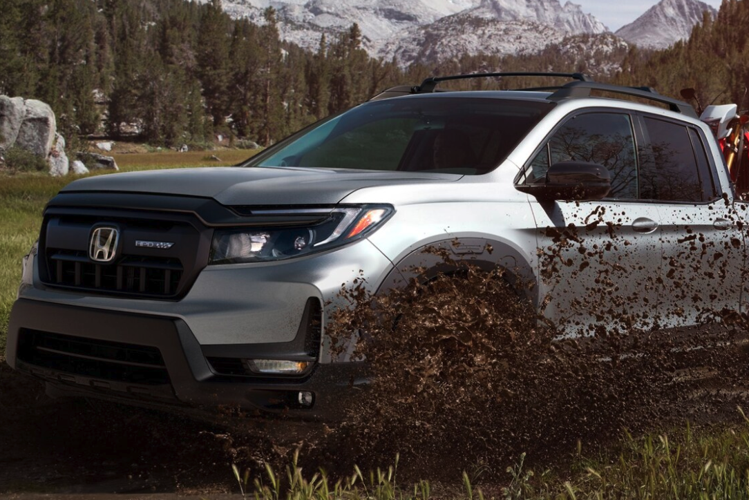 A silver Honda Ridgeline pickup truck with a roof rack splashes mud forward while driving through a grassy, wooded area with rocky mountains in the background.