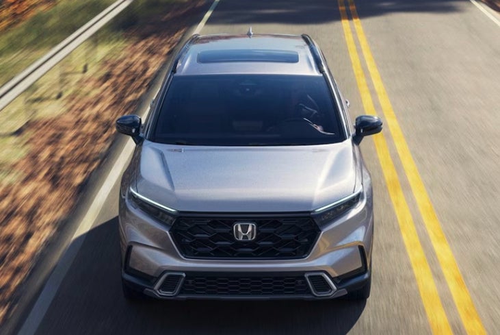 A silver Honda CR-V crossover is seen from a high-angle view, driving down an asphalt road lined with trees and double yellow lines.