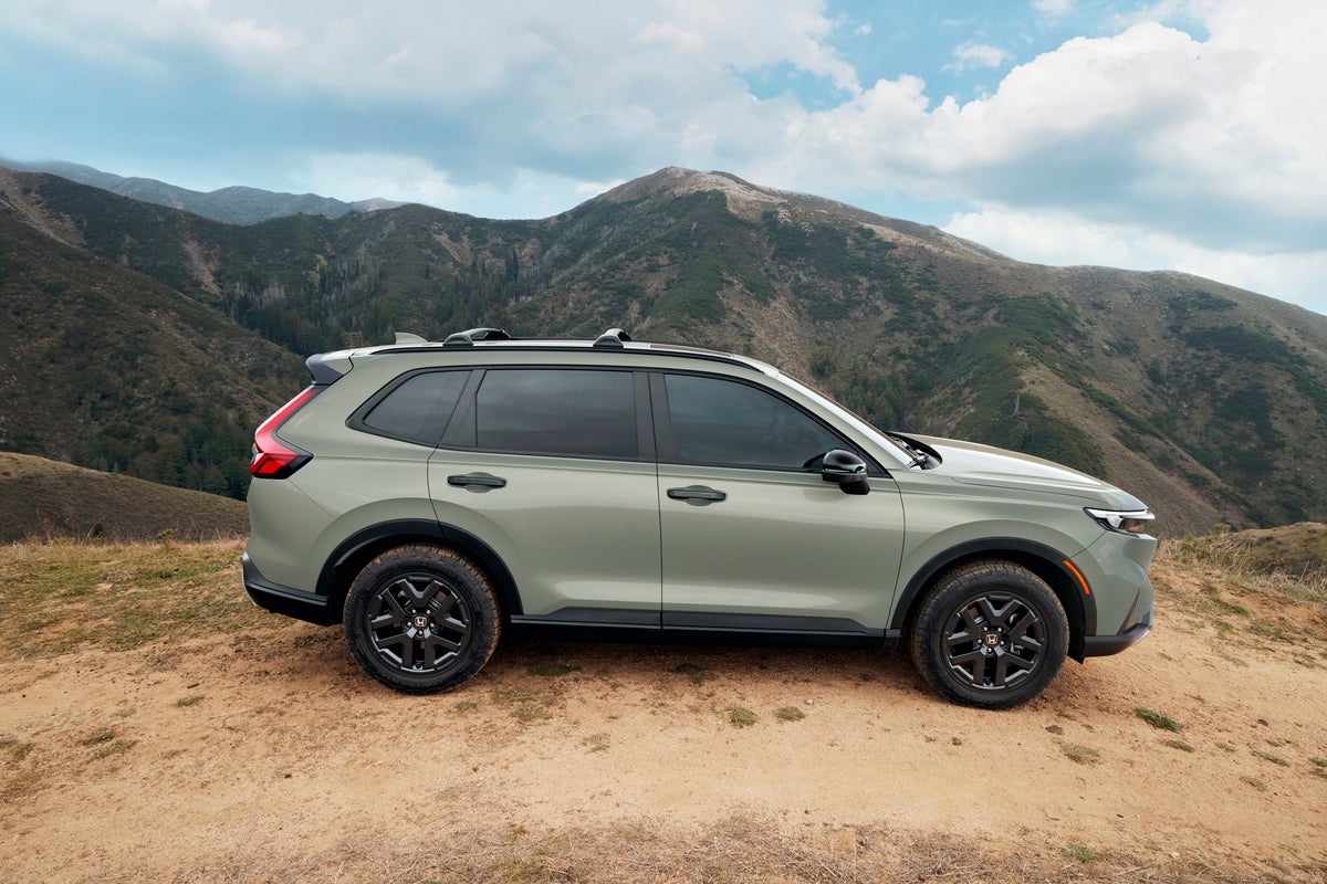 A light green SUV is parked on a dirt overlook with rugged, tree-covered mountains in the background. The vehicle is shown in a full side profile, highlighting its black wheels and roof rails under a partly cloudy sky.