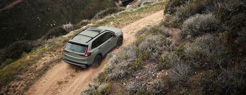 A light green SUV drives along a narrow dirt road on a rugged hillside, surrounded by dry shrubs and rocky terrain. The vehicle is viewed from above and behind as it moves upward along the winding path.