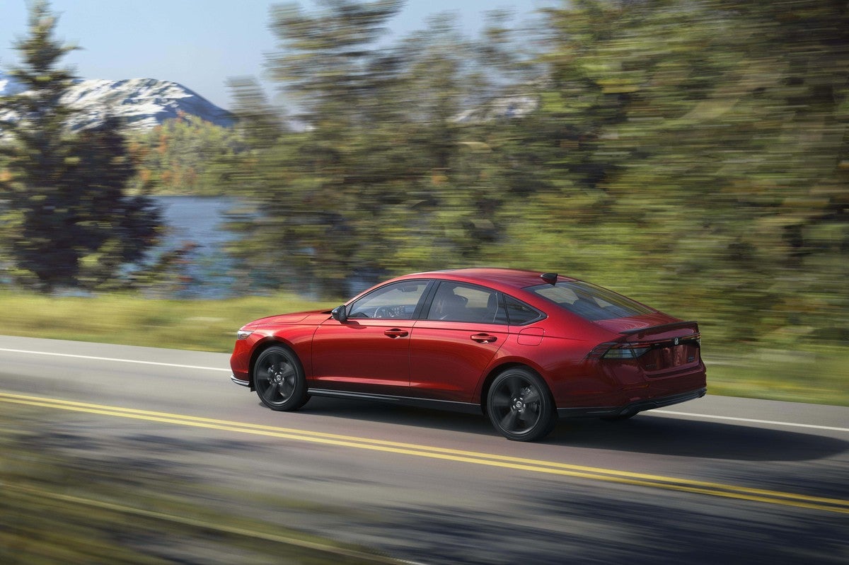 A red 2026 Accord in Phoenix drives away from the viewer to the left on a two-lane country road with a lake and mountains in the background.