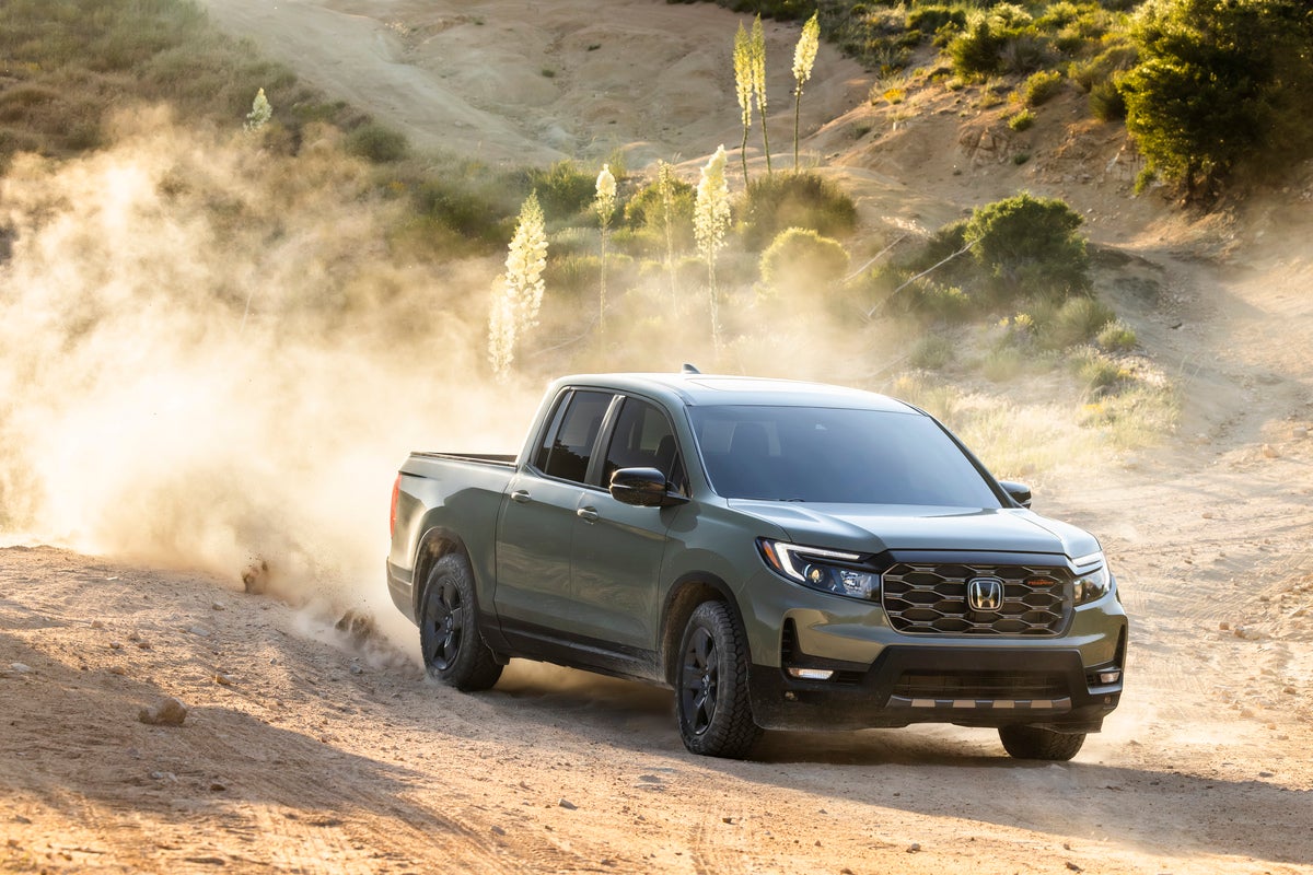 An ash green metallic 2026 Honda Ridgeline Trailsport drives towards the viewer to the right down a dusty desert slope, kicking up dust behind it.