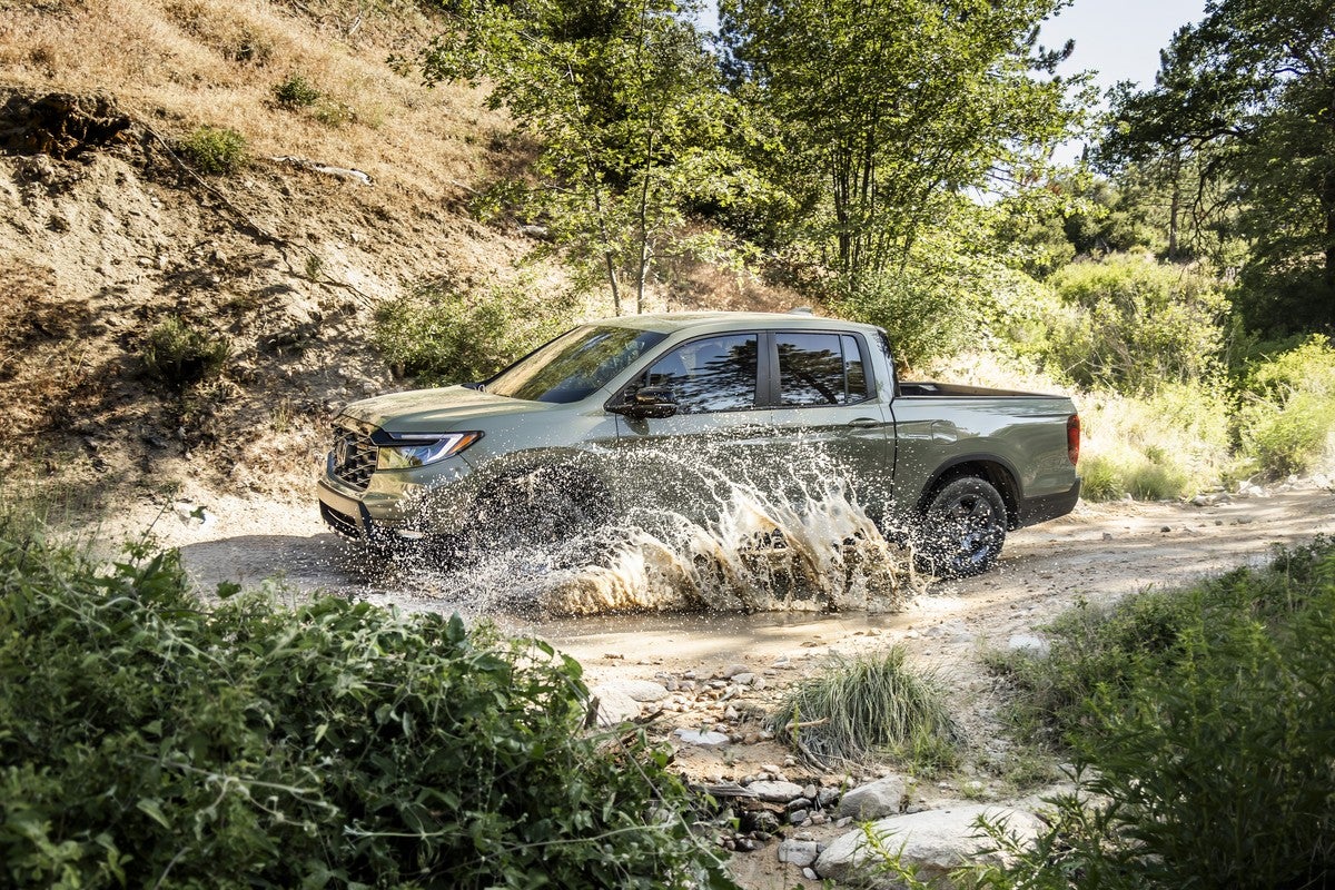 An ash green metallic 2026 Honda Ridgeline trailsport drives to the left on a dusty road through a puddle of dirty water, splashing the water towards the viewer.