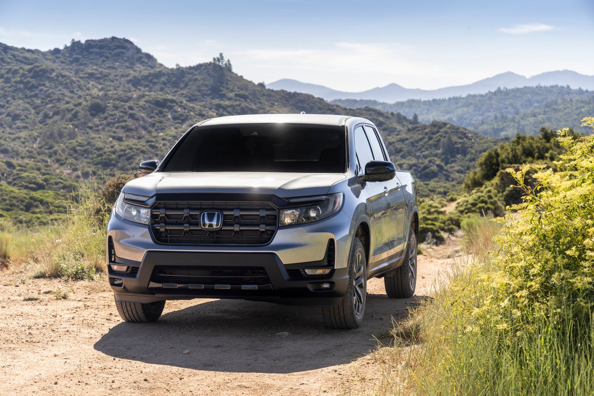 A solar silver 2026 Honda Ridgeline Sport faces the viewer on a rural dirt road with arid hills in the background.