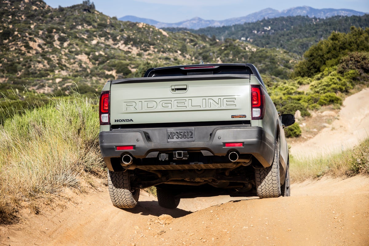An ash green metallic 2026 Honda Ridgeline Trailsport drives away from the viewer down a bumpy dirt trail with arid hills in the background.