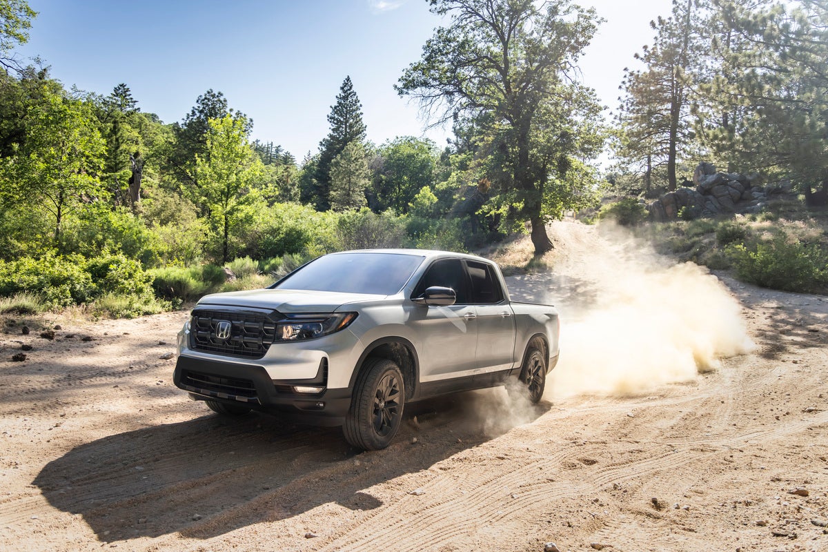 A solar silver metallic 2026 Honda Ridgeline Sport drives towards the viewer to the left up a dusty trail, kicking dust behind it.