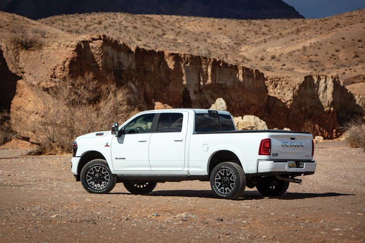 A white Ram heavy-duty pickup truck is parked on a dusty plain with tall, dry, rocky cliffs in the background.