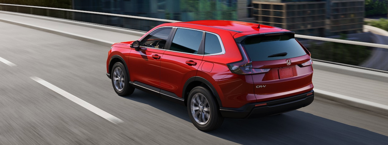 A red Honda CR-V SUV drives away on a paved road, with blurred city buildings in the background.