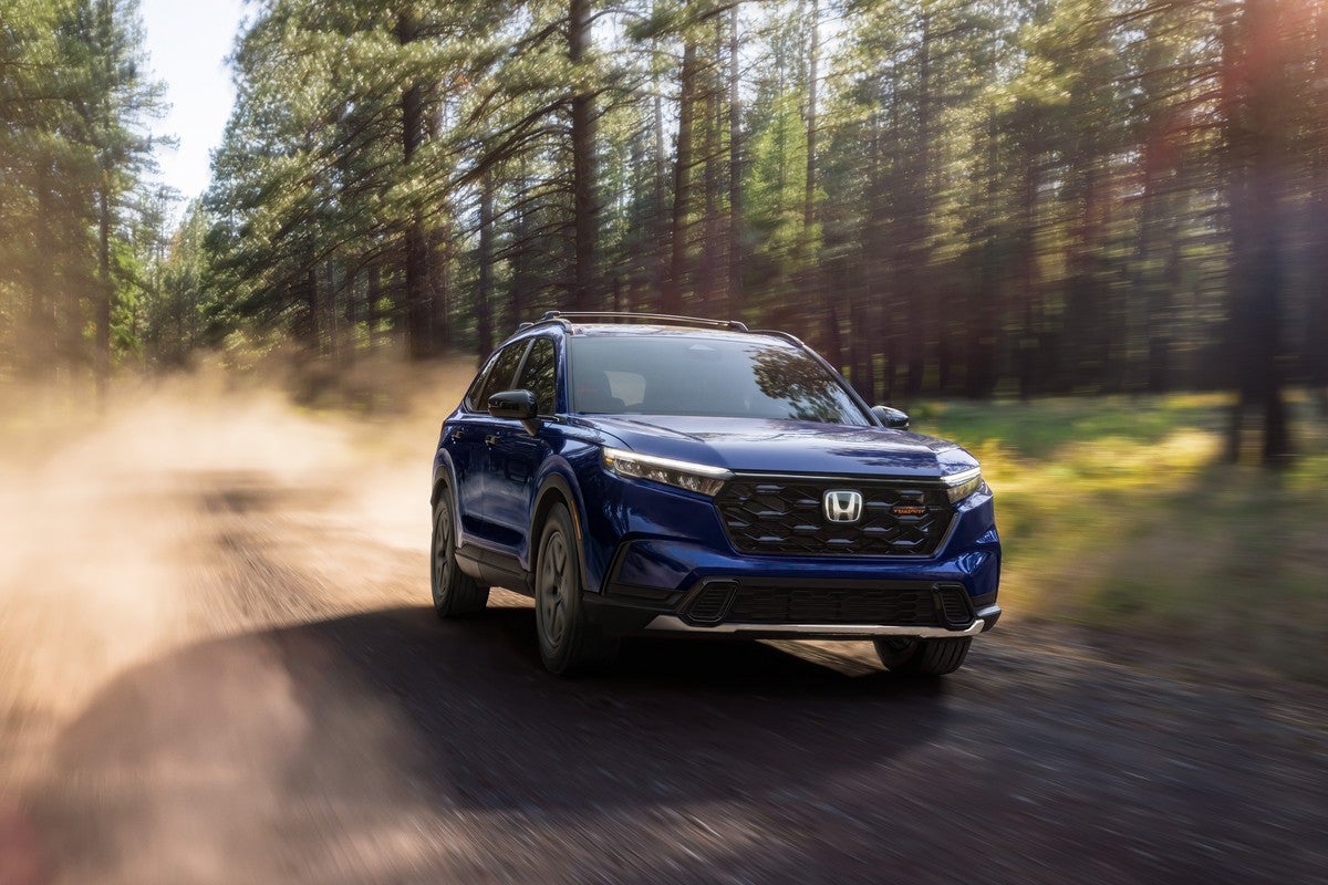 A blue 2026 Honda CRV in Phoenix drives towards the viewer to the right on a dirt road through a forest.