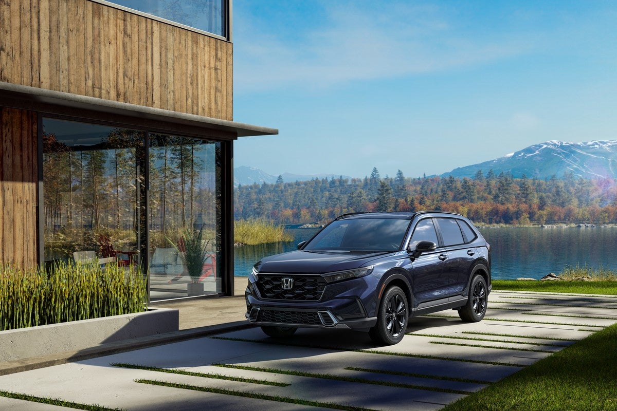 A blue certified preowned Honda CRV in Phoenix is parked facing the viewer to the left ina driveway next to a modern house with a lake and tree-covered mountains in the background.
