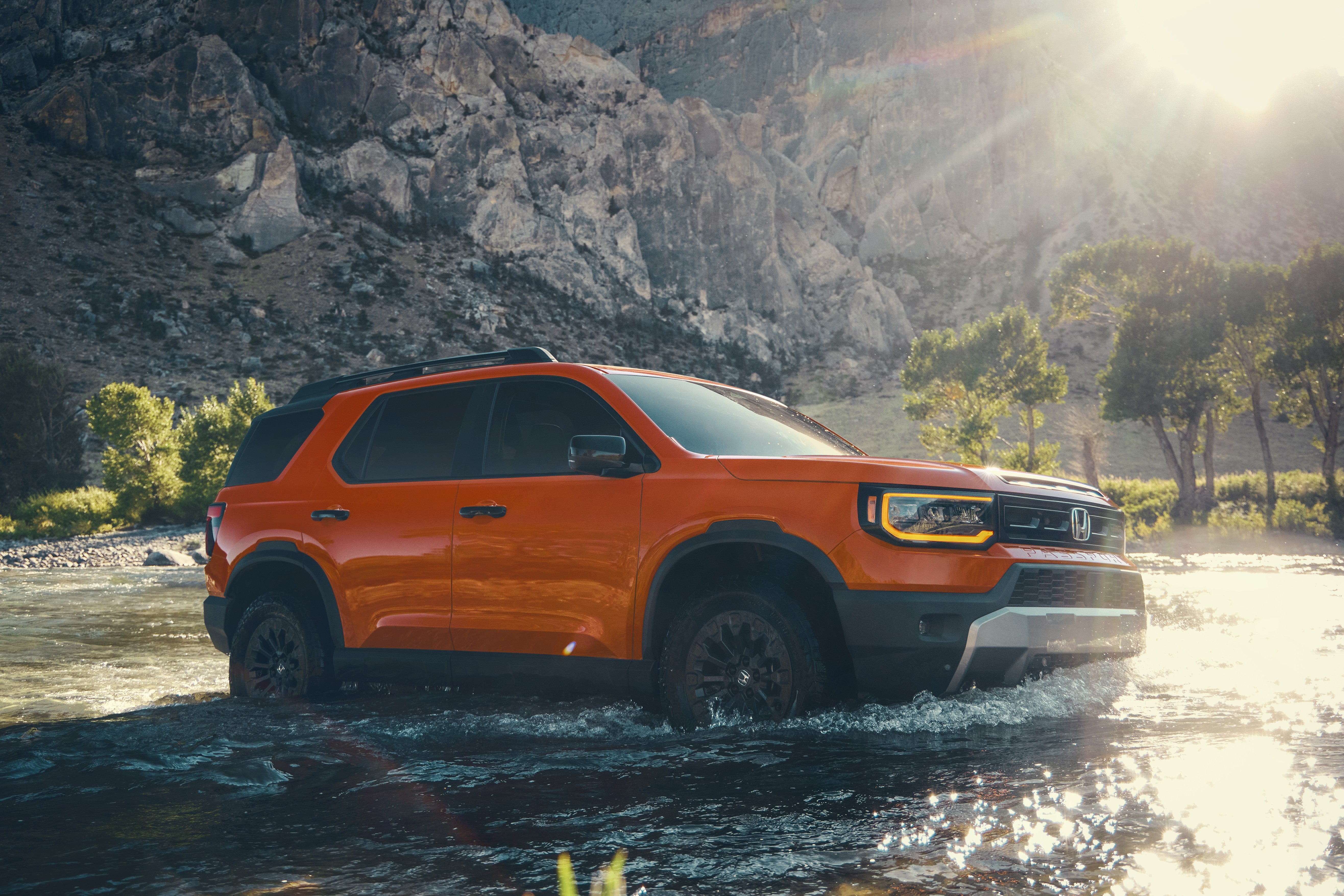 An orange new Honda Passport for sale in Phoenix drives to the right through a shallow river with a the rocky face of a large cliff in the background.