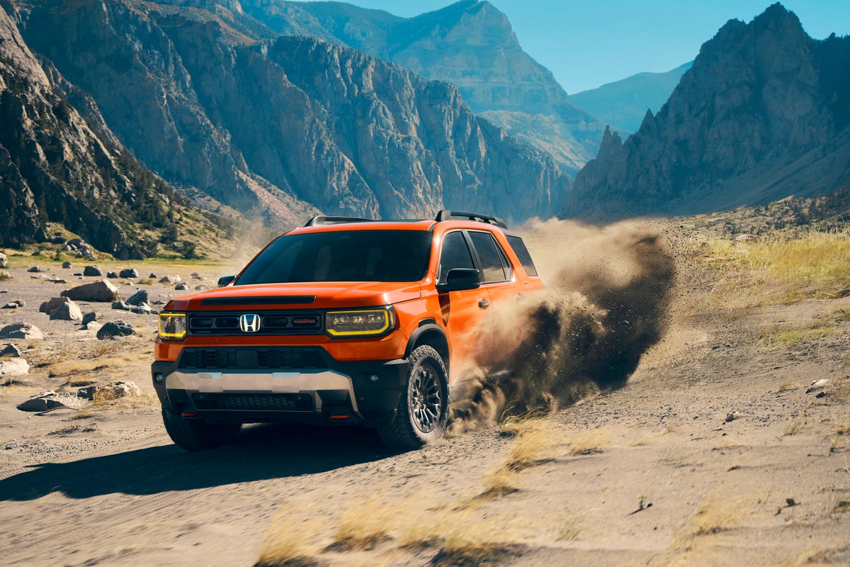An orange new Honda Passport in Phoenix drives towards the viewer to the right while kicking up a wave of dirt in the bottom of a large valley surrounded by massive cliffs.