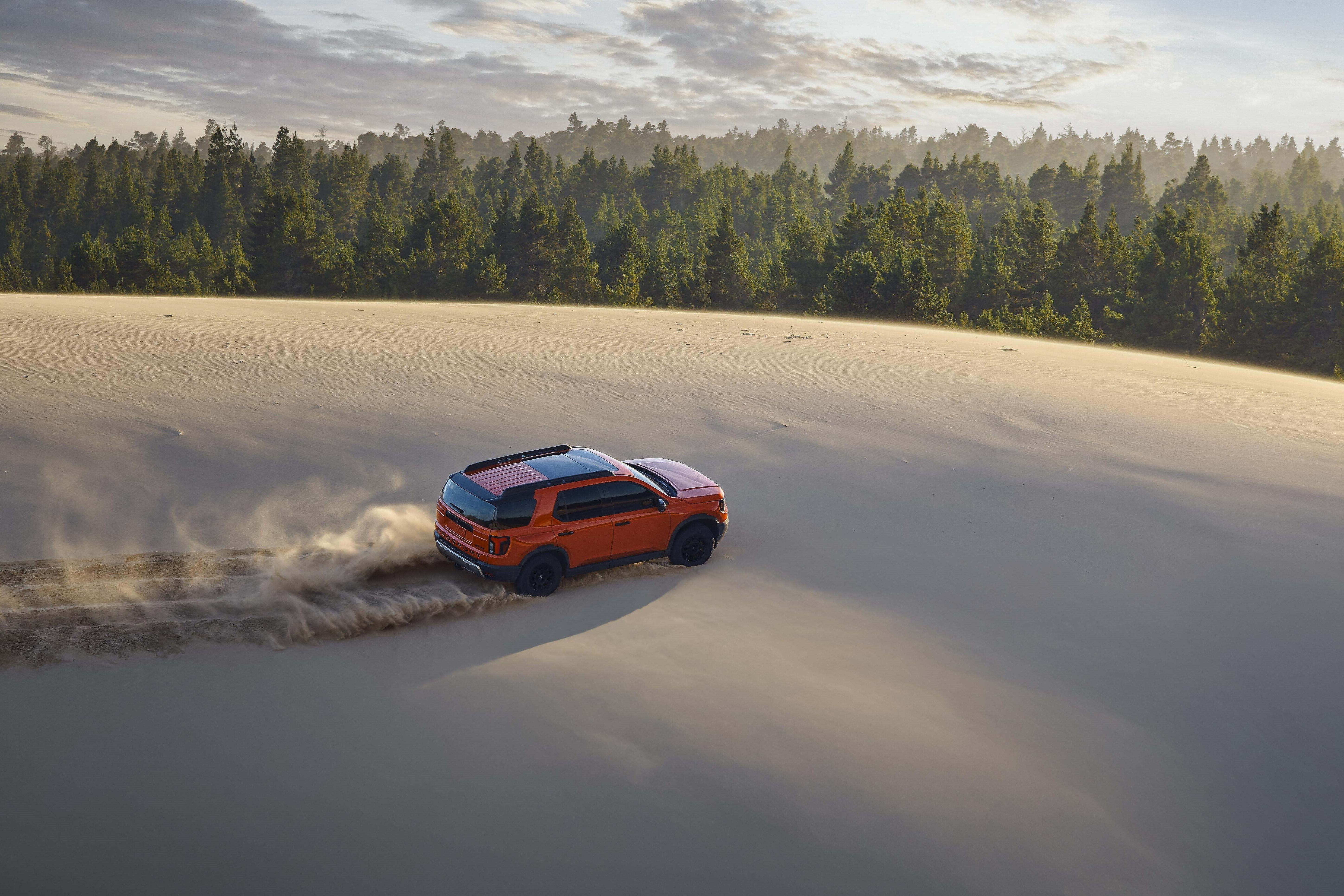 An orange used Honda Passport for sale in Phoenix drives away from the viewer to the right seen from a high angle through the a desert kicking up sand.