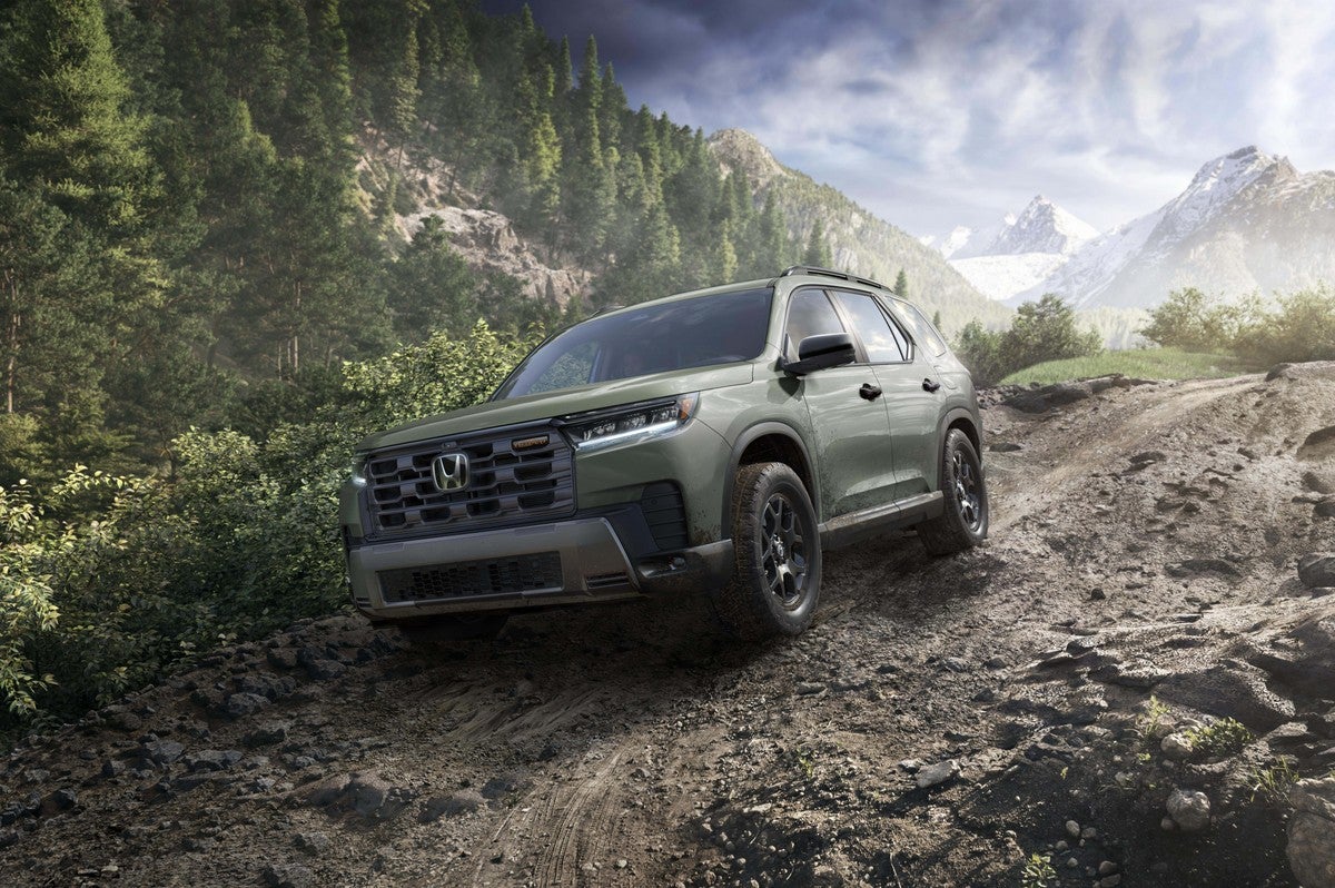 A green 2026 Honda Pilot in Phoenix drives towards the viewer to the left down a steep mountain dirt road with a snowy peak in the background.