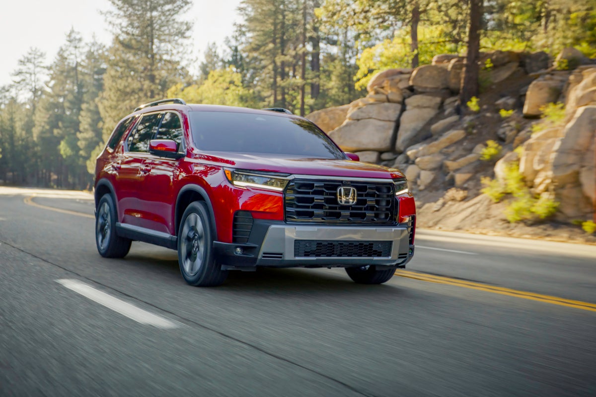 A red certified preowned Honda Pilot in Phoenix drives towards the viewer to the right on a rural road amongst trees.