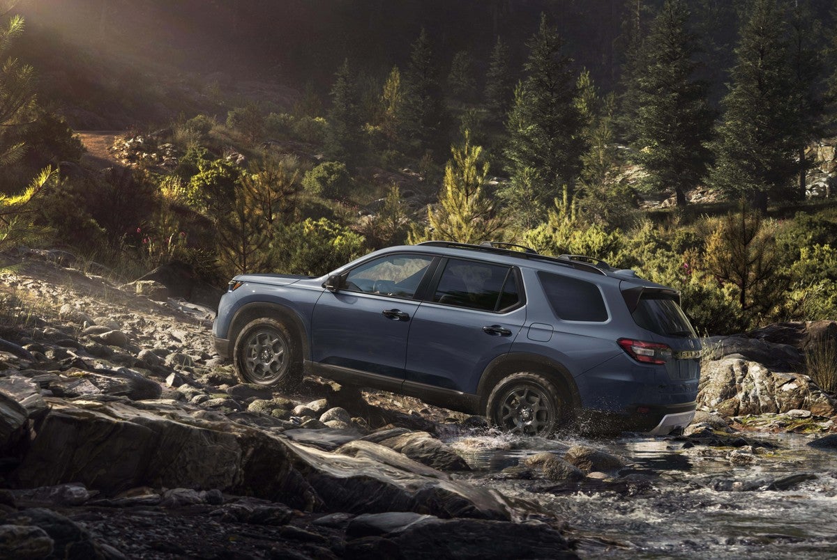 A blue used Honda Pilot in Phoenix drives away from the viewer to the left up a steep slope of boulders on a tree-covered mountainside.
