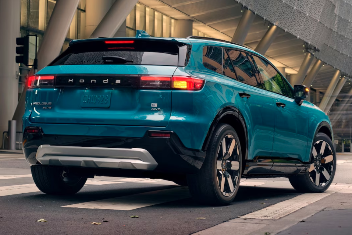 A three-quarters rear view of a turquoise Honda Prologue electric SUV. The vehicle is on a city street, with a modern building in the background. The 'HONDA' lettering is prominent across the back tailgate, and the vehicle has a two-tone paint scheme with a black lower body and a silver rear bumper accent.