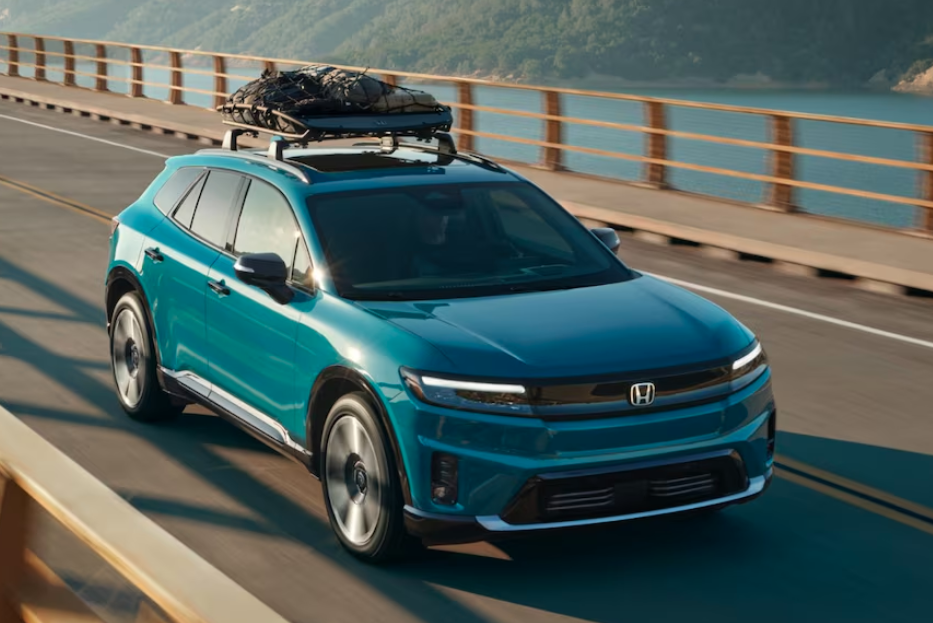 Blue Honda SUV driving along a coastal highway with ocean on one side and rocky mountains on the other, under a bright blue sky.