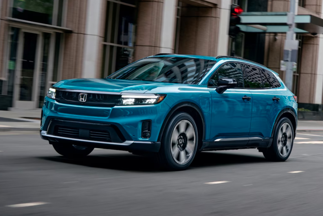A three-quarters front view of a blue Honda Prologue electric SUV driving on a city street. The vehicle is in motion with a slightly blurred background of buildings, and its wheels are also blurred, indicating movement.
